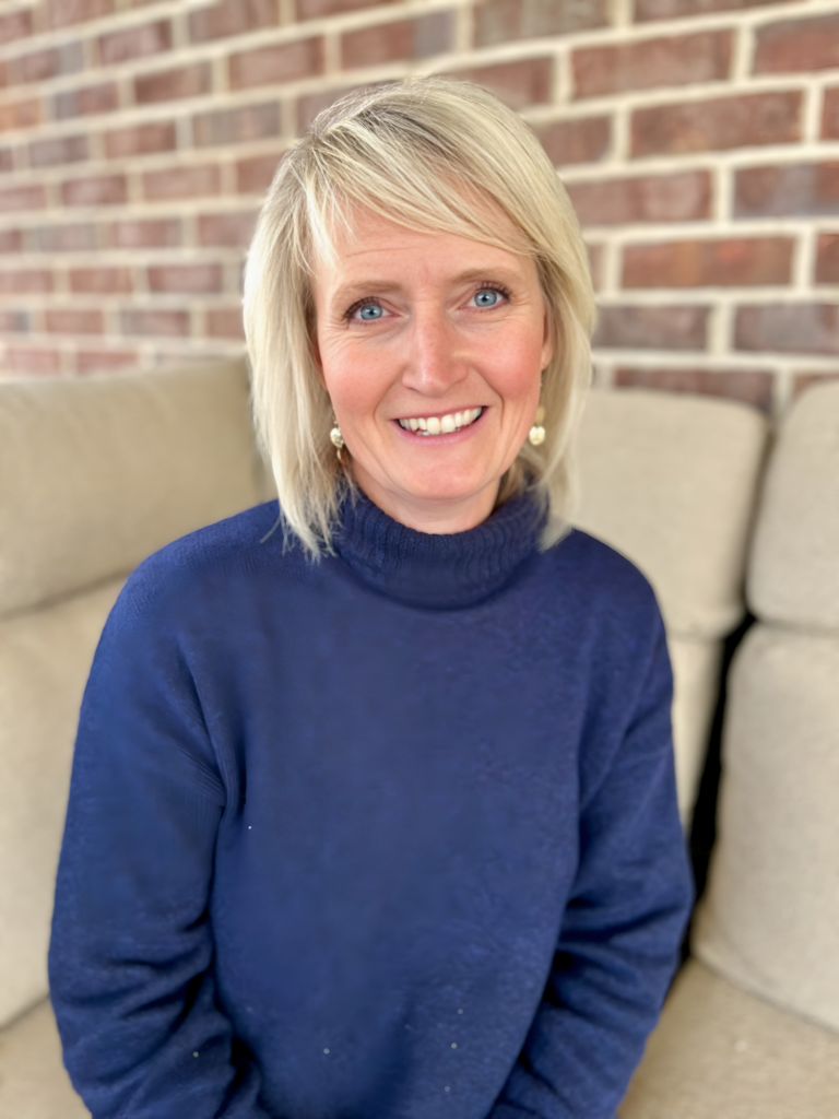 Joy Christensen smiling in a blue sweater, sitting against a brick wall backdrop